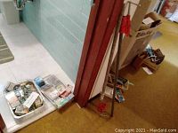 Photo showing 2 red-handled bar clamps standing against the wall and trays on floor with hardware items.