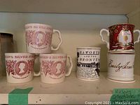 Shelf display of six collectible commemorative mugs including three Queen's Silver Jubilee mugs, Haworth and the Brontes mug, Emily James mug, and a Golden Jubilee mug.