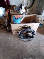Box containing coiled black electrical cord, blue foam pad in packaging, and other camping supplies.