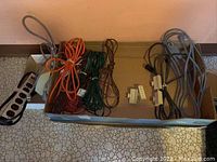 Lot of various extension cords and power bars in an open cardboard box on tiled floor, showing black, orange, green and brown cords and white power adapters.
