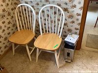 Photo showing two white painted wooden kitchen chairs with natural pine wood seats. The seats have visible chips in the finish. Next to the chairs is a small stainless steel Simple Human trash can with a foot pedal and sensor, topped with a box of trash bin liners.