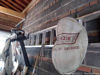 Photo of the left side view showing the folded 20ft aluminum extension ladder hanging horizontally on a brick wall, with the Toro Ultra leaf blower handle and bag with 'Mackenzie Recreational Vehicles' logo visible hanging below.