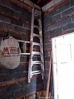 Full-height photo of an aluminum ladder leaning against a brick wall in a garage or workshop space. The ladder shows wear and age.