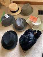 Seven vintage men's hats displayed on carpet, including Dunns UK Homburg, various felt hats, cream outdoor hat, and black fur hat with ear flaps