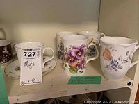 Photo showing four floral mugs and one teacup with saucer on shelf.