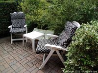 Two white folding resin patio chairs with black and white geometric patterned cushions and one white resin patio table on brick patio surrounded by greenery.