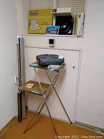 Keystone Gravity 1200 projector on two-tier folding stand with wood-pattern shelves and chrome legs, alongside folded projector screen parts leaning on wall, and boxed Kenzo slide sorter on shelf above.