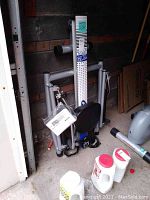 Photo of the folded Stamina 1215 Orbital Rower machine in a garage, showing the black seat, pedals with straps, and the vertical support panel with branding and instructions.