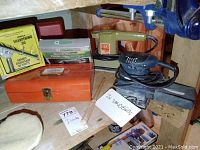 Wide shot showing two sanders, toolbox, drill sharpening jig box, glue gun box, and a polishing pad on the workbench.