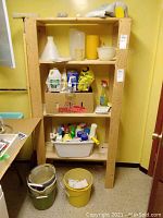 Front view of the wooden shelving unit with several objects on shelves and buckets on floor, yellow painted walls in the background.