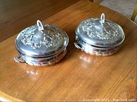 Two ornate silver plated covered casserole dishes with floral embossed lids and loop handles, resting on wooden table
