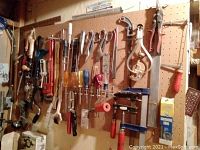 Photo showing assortment of hand tools arranged on pegboard including screwdrivers, hand drills, saws, clamps, scissors, and pipe wrench.