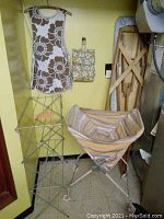 Photo showing a wire foldable clothes drying rack, fabric laundry cart with striped fabric, wooden ironing board with floral cover, fabric clothes pin bag, and wooden pants stretchers hung on the wall.