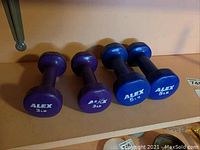 Four hand weights displayed on a shelf: two purple 3 lb dumbbells and two blue 5 lb dumbbells, all labeled ALEX.