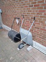 Metal garden wheelbarrow and metal garden roller leaning against a brick wall on a paved surface. Both show signs of pitting and rust.