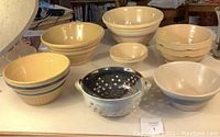 Photo of a set composed of six vintage yellow-beige pottery bowls with varied colored stripes and different sizes, a small white bowl, and a blue-gray pottery colander with drainage holes and side handles, all showing signs of age including chips and cracks.