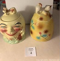 Two vintage cookie jars on table: Man in the Moon face jar with kitten on lid and side; and yellow beehive jar with floral decoration and kitten on top.