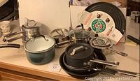 View of multiple assorted pots and pans on counter including saucepans with lids and frying pans from Cuisinart, Calphalon, and All-Clad.