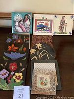 Photo of eight decorative art tiles displayed on a table, showing various styles and colorful designs including figurative, floral, and landscape themes.