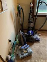 Overall photo showing a group of vacuum cleaners and cleaning supplies leaning against a wall in a carpeted room.