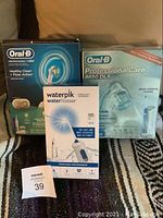 Lot of two unopened Oral-B electric toothbrush boxes and one Waterpik cordless water flosser box placed on fabric background.