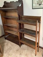Photo showing the large wooden bookcase with decorative top and the small mission style bookcase with three shelves side by side on carpeted floor.