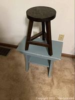 Dark brown round wooden stool on top of light blue shabby chic table, beige carpet background