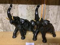 Front view of the two black cast metal elephant figurines with trunks raised, standing on a flat surface against a stone or concrete wall background.