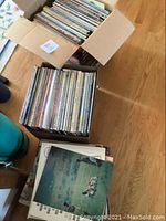 Two open boxes filled with vinyl records, viewed from above on wooden floor. Many album covers and boxed sets are visible showing classical and jazz themes.