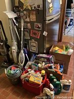 Photo of vacuum cleaners and cleaning supplies in buckets and boxes near fridge.