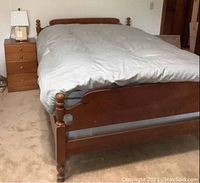 Full view of the full size maple wood bed frame with made bed and matching nightstand showing lamp on top