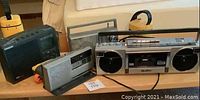 Photo showing 4 assorted radios and tape players placed on a wooden surface in front of a beige couch. Items include a silver boombox, a silver portable cassette player, a black digital radio, and a black portable antenna radio.