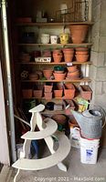 Shelf with multiple sizes and styles of terracotta pots and saucers along with some plastic and ceramic pots, a painted wooden plant stand in front and various garden buckets and watering cans on floor below.
