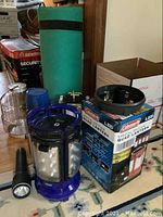 Photo showing camping gear items on floor including Coleman LED Quad Lantern in box, green rolled sleeping pad, thermos, two flashlights, blue cup, glass lantern globe, and other camping goods.