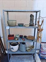 Photo shows overall lot on metal shelving including various glazed pots, garden statue, metal trivet, watering can, and bamboo wind chime.