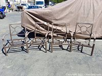 Four brown metal outdoor chairs arranged side by side, showing full frames and armrests, with no cushions.