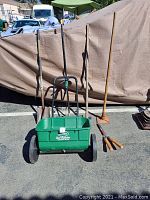 Six garden tools arranged upright and leaning against a covered object outdoors, including broadcast spreader, cultivator, leaf rake, and three hand tools.