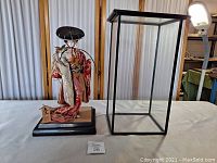 Side-by-side photo of the Geisha doll mounted on base with glass display case frame beside it showing scale and condition.