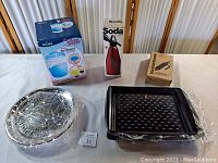 All five new kitchen accessory items shown on table: Rival Ice Shaver box, Soda Spritzer box, Pampered Chef Accent Decorator box, four slip-proof serving trays wrapped in plastic, and a black stovetop grill wrapped in plastic.