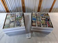 Two white corrugated full-card storage boxes on table, each packed with unsorted cards topped by loose stacks of football and baseball cards