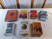 Front view of seven books laid out on a white surface, showing cover designs and titles from George R.R. Martin's "Game of Thrones" series.