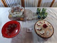 Photo of vintage kitchenware displayed including red glass bowl, ceramic pie keeper, green glass cups, clear glass serving dish in metal holder, and metal stand