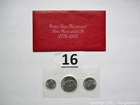Photo of red holder with set of three coins in sealed plastic sleeve placed below it, lot number "16" present.