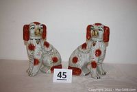 Pair of seated ceramic spaniel dogs, creamy white with reddish-brown spots, chain and locket detail, against white background with lot number sign in front.