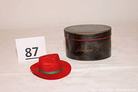 Photo of the miniature red fedora hat next to its original black round Stetson hat box with gold logo and red trim.