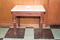 Front view of small rectangular table with white marble top and dark walnut wooden base featuring turned legs, placed against wood panel wall on checkered floor.
