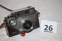 Front and left side view of vintage Argus Autronic 35 camera in partial leather case with camera strap and visible lens and shutter button. Number card '26' and red dice placed in front.