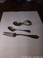 Photo showing the three silver utensils: a baby spoon with folded handle, a small spoon, and a fork all tarnished and aged.