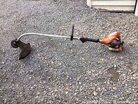 Full view of the entire Echo GT-225 gas trimmer laid on gravel, showing the curved metal shaft, orange engine, black handle, and cutting guard.
