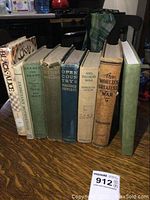 Seven antique and vintage hardcover books standing upright showing spine titles with varying shades of green, beige, brown, and white covers.
